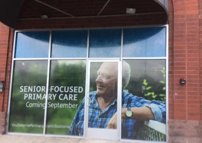 A multi window print on an exterior wall with entry door. Shows a senior man with text that reads, "Senior Focused Primary Care Coming September... YouDeservePartners.com/Gastonia".