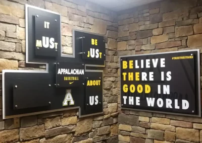 Wall art present on two intersecting stone masonry interior walls. Mostly consisting of text divided between several frames. Messaging reads "Believe there is good in the world". Another frame includes both the App State Basketball team logo and workmark paired with the Nike Check logo.