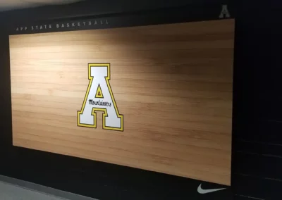 Wall mural for Appalachian State Mountaineers basketball team on a dark blue wall. Logo printed with a large Letter "A". The word "Mountaineers" is written inside the middle bar of the letter "A" in cursive typeface. The logo is printed on a wood plank background spanning a large portion of the wall. Above reads "App State Basketball" on the left and a small version of the same logo position on the right. At the bottom right is a Nike logo.