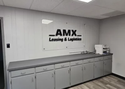 Logo with wordmark printed on a large pane of glass hanging installed on an interior wall above cabinets and countertop. Logo icon is big bold text reading "AMX". Below is the tagline reading "Leasing & Logistics".