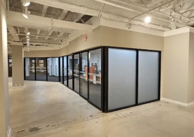 An office space under construction. Glass doors and floor to ceiling windows are seen with two sets of double lines running horizontally through the vertical center of each window. Two windows are frosted without lines.