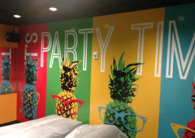 An interior office mural with large vertical stripes colored in red green yellow blue and orange. A print of a pineapple with glasses sits on each vertical stripe. Large text reads, "It's Party Time".